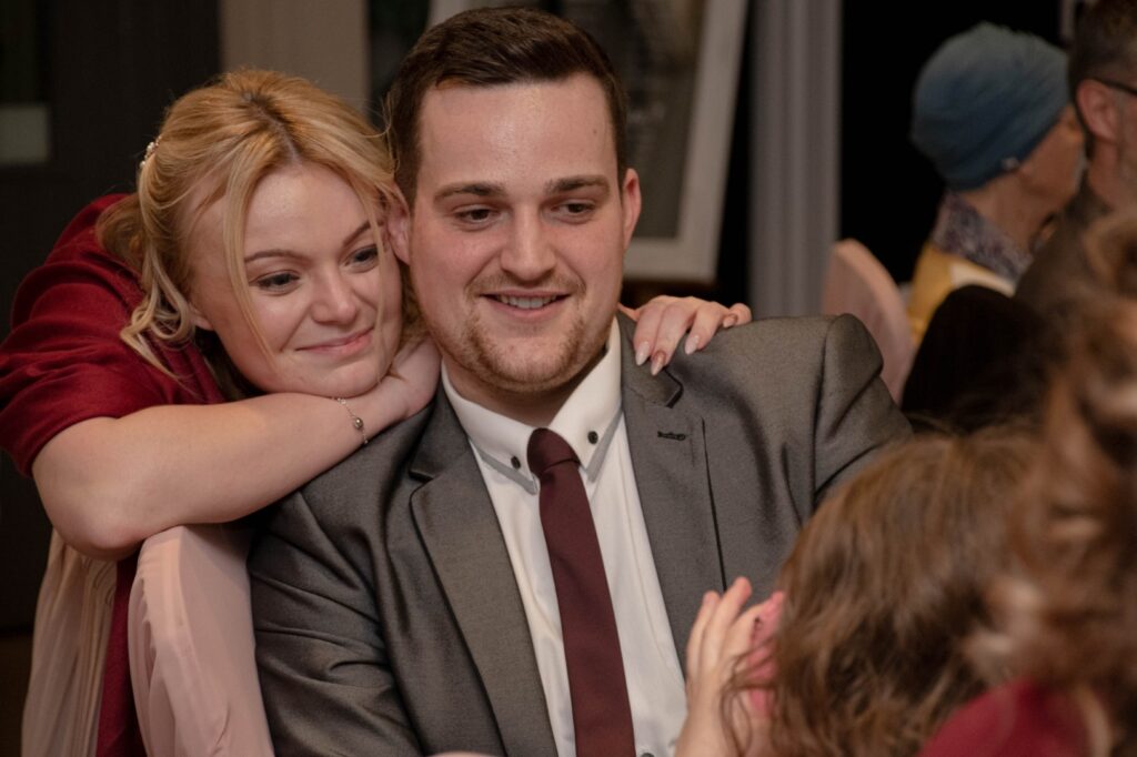 A smiling man in a suit and a woman in a red dress embrace warmly at a social gathering, surrounded by softly blurred guests in the background.