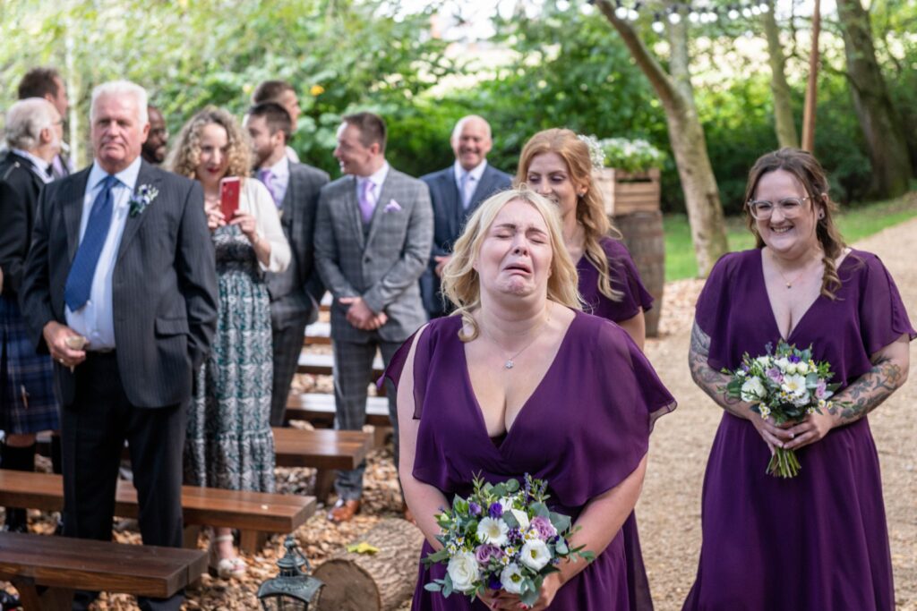 A woman in a purple dress expresses strong emotion while holding a bouquet, surrounded by guests in formal attire during an outdoor wedding ceremony.