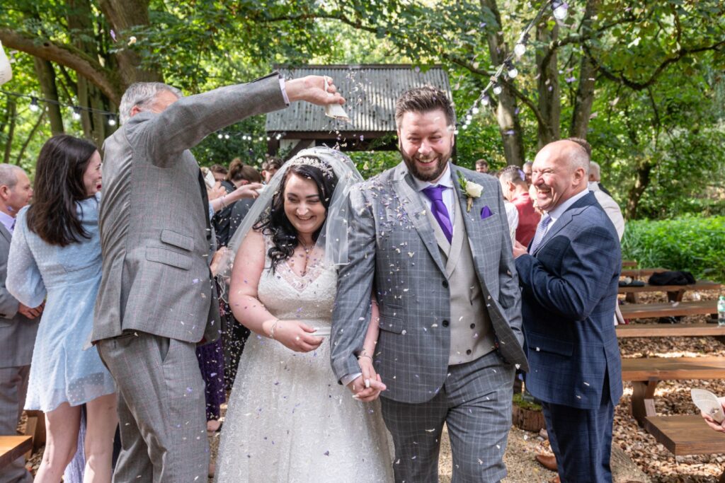 A joyful couple walks together, surrounded by guests showering them with colorful confetti in a beautiful outdoor setting.