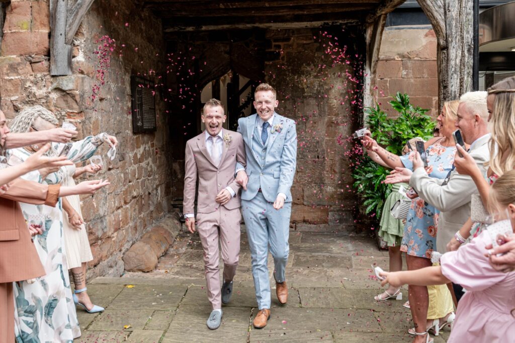 Two grooms happily walk through a crowd celebrating their marriage, with rose petals being thrown in the air.