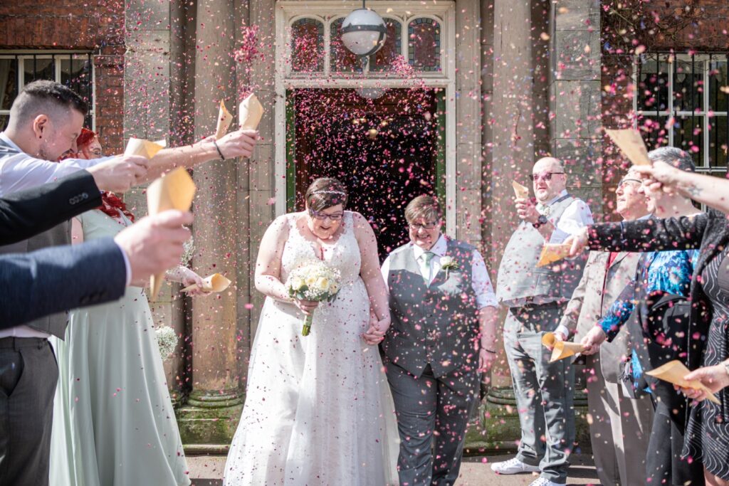A newly married couple walks through a shower of colorful confetti, surrounded by smiling guests celebrating their union in front of a historic building.