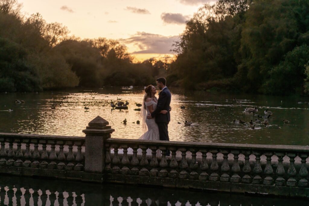 A couple in formal attire gazes lovingly at each other on a stone bridge over a tranquil pond, surrounded by ducks and a scenic sunset backdrop.