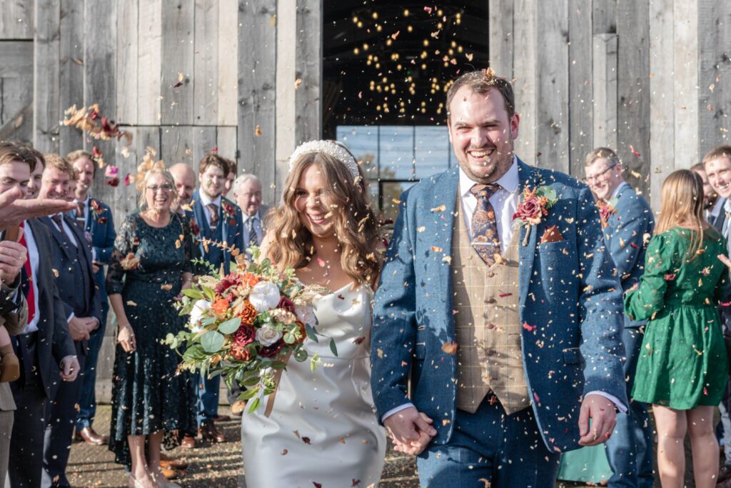 A newlywed couple walks hand in hand, smiling and surrounded by confetti as they exit a barn, with guests cheering in the background.