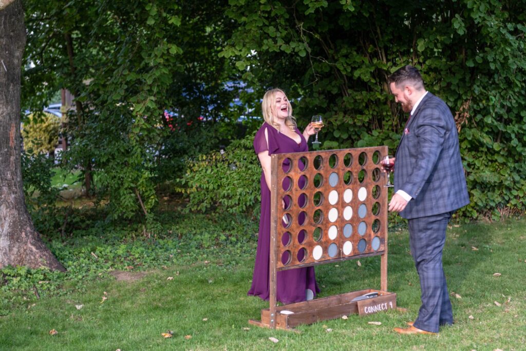 A woman in a purple dress and a man in a plaid suit playing Connect Four outdoors, surrounded by greenery, with drinks in hand and joyful expressions.