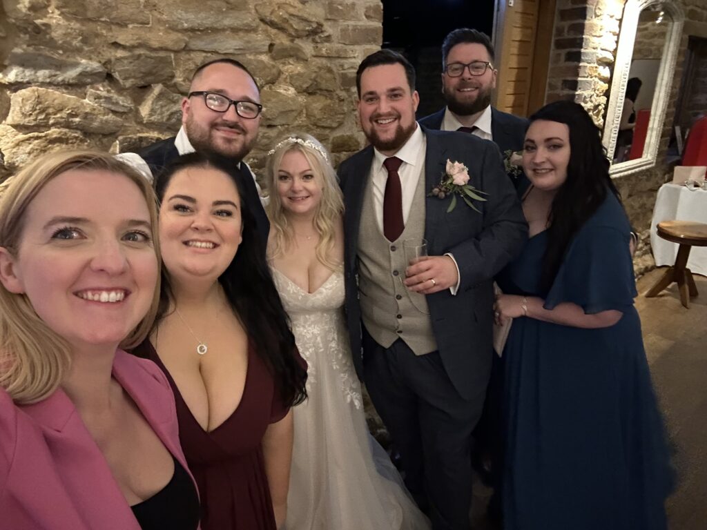 A group of friends and family posing together at a wedding reception, with the bride in a wedding gown and joyful expressions all around.