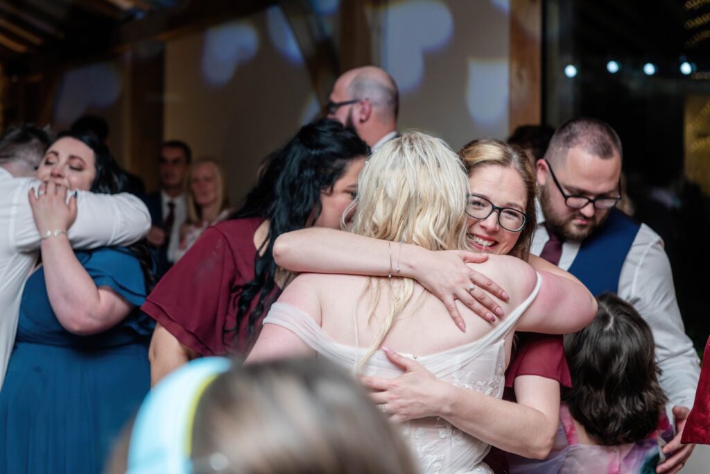 Two women embrace joyfully amidst a festive wedding atmosphere, surrounded by friends and family.