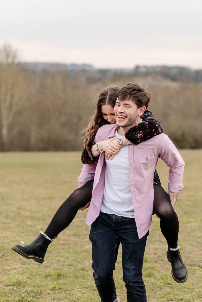 A man carries a woman on his back, both laughing, while enjoying a playful moment outdoors in a grassy field.
