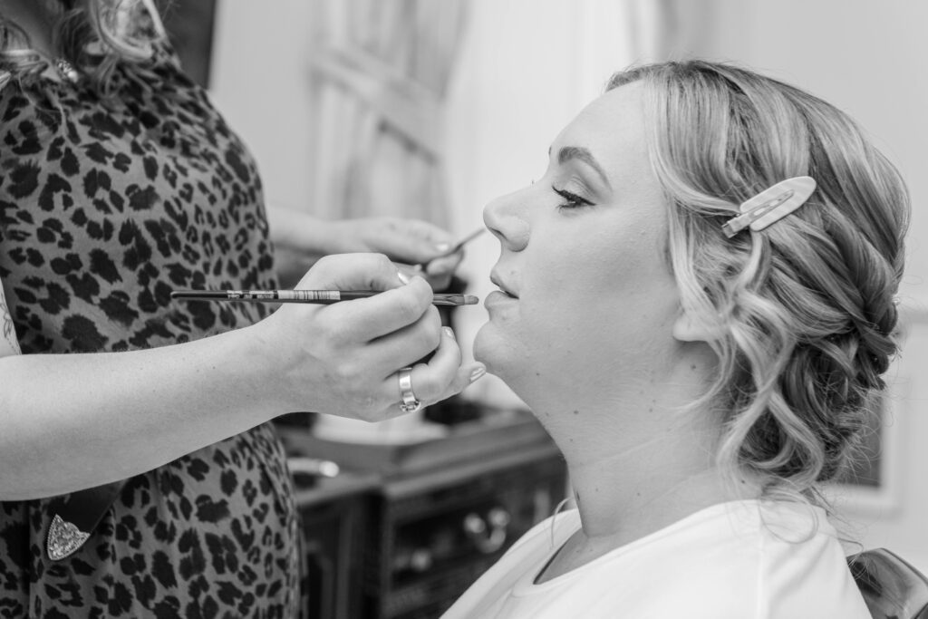 A close-up of a woman receiving makeup application, with a makeup artist working on her lips, set in a softly lit environment.