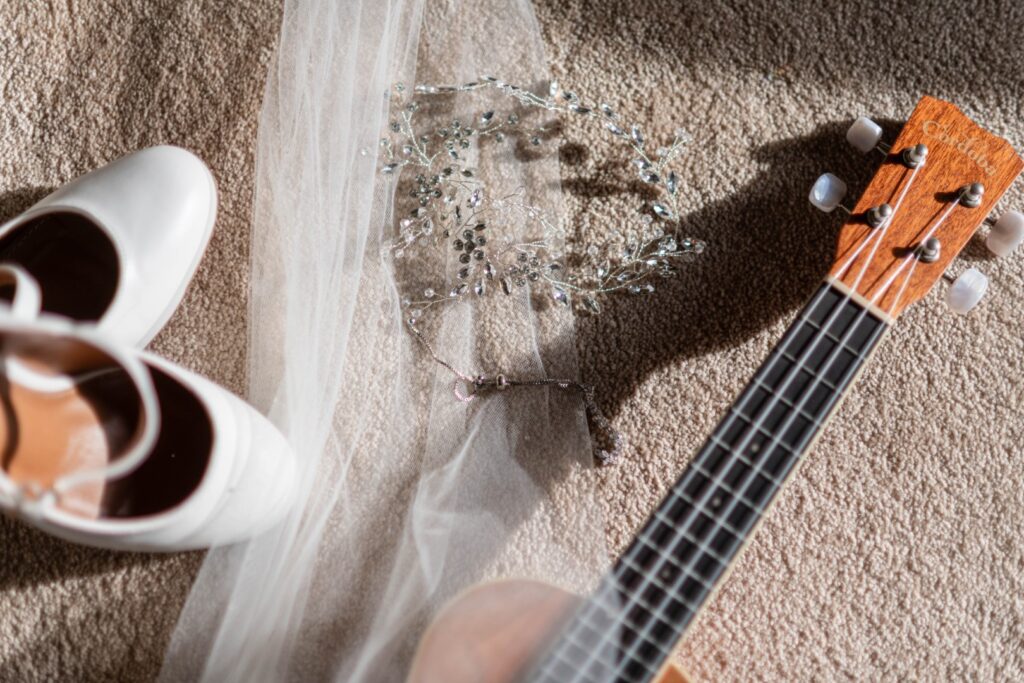 A collection of wedding accessories including white shoes, a delicate veil, a sparkling hairpiece, and a ukulele, arranged on a textured surface.