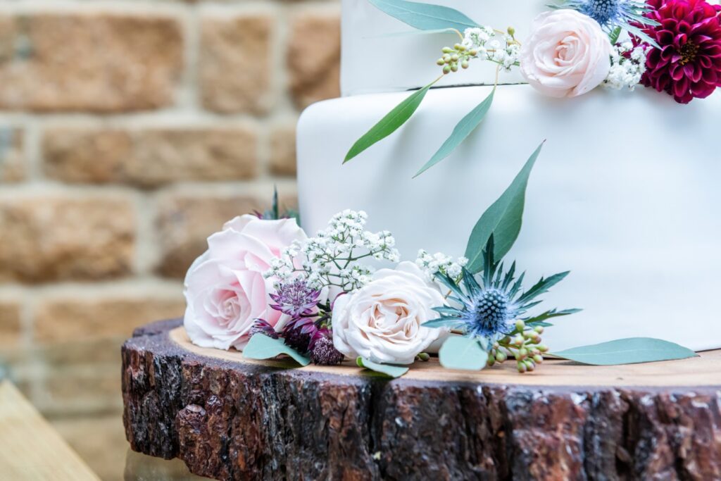 A beautifully decorated wedding cake featuring pastel roses, greenery, and colorful flowers on a rustic wooden base, set against a stone background.