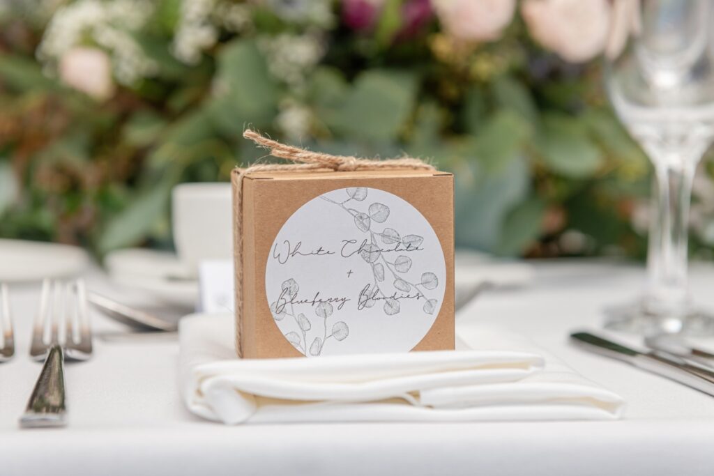 A decorative gift box labeled 'White Chocolate + Blueberry Blondies' sits on a neatly arranged table with silverware and a background of greenery and floral arrangements.