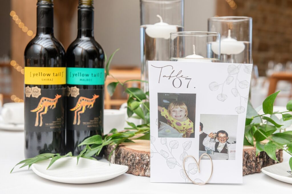 A stylish table setting featuring bottles of Yellow Tail Shiraz and Malbec, a table number card with childhood photographs, and decorative greenery.