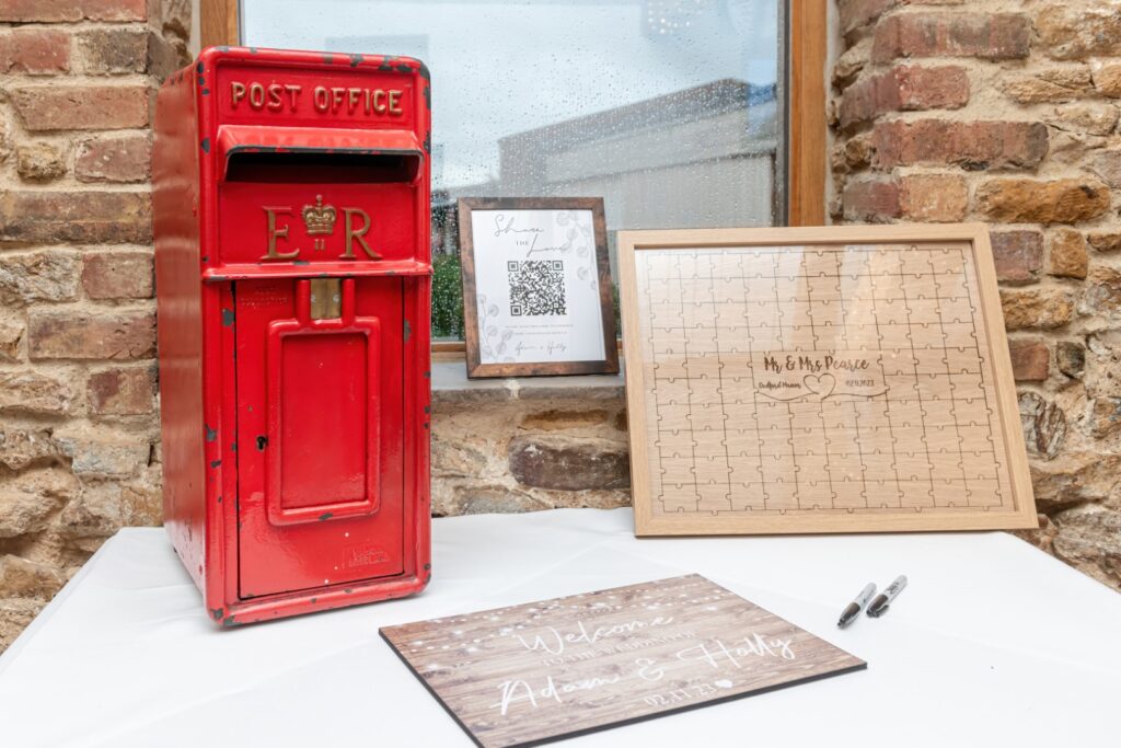 A vintage red post box beside a welcome sign and a wedding puzzle guest book, set against a brick wall.