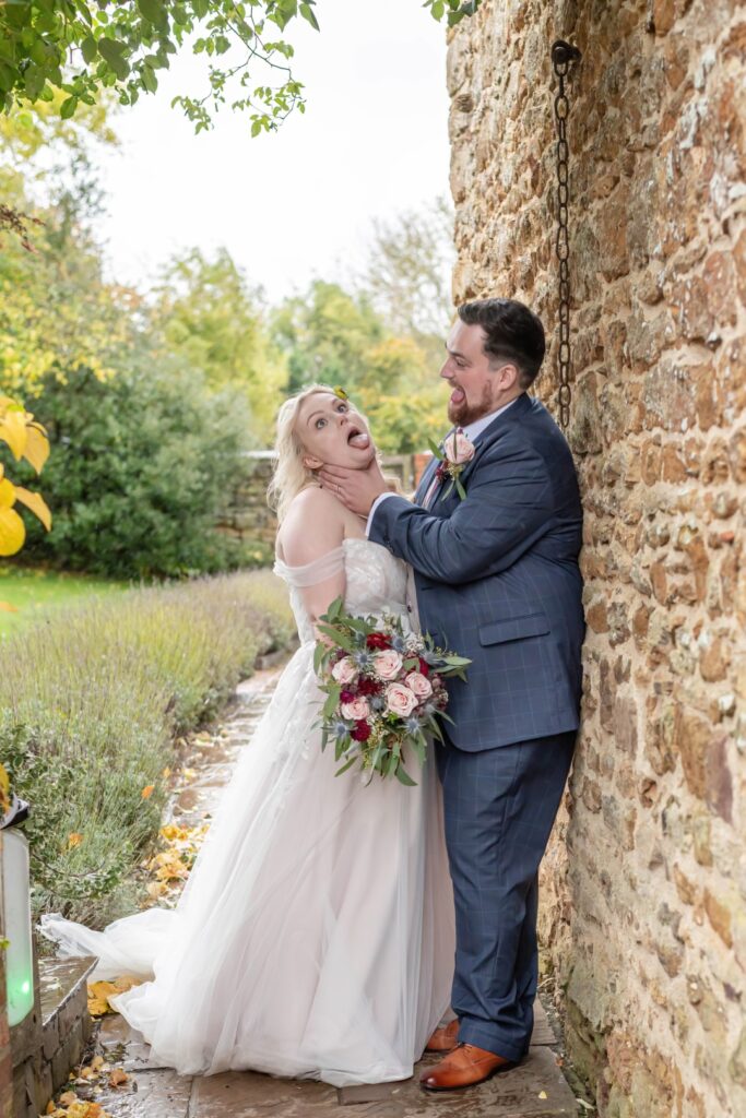 A couple in wedding attire sharing a playful moment, with the bride making a silly face while the groom smiles, surrounded by greenery and stone walls.