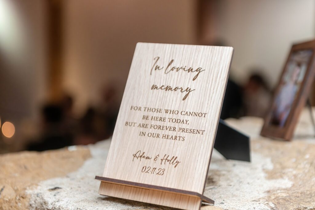 A wooden tribute sign commemorating those who cannot be present, featuring elegant text and heartfelt dedication for Adam and Holly dated 02.11.23.
