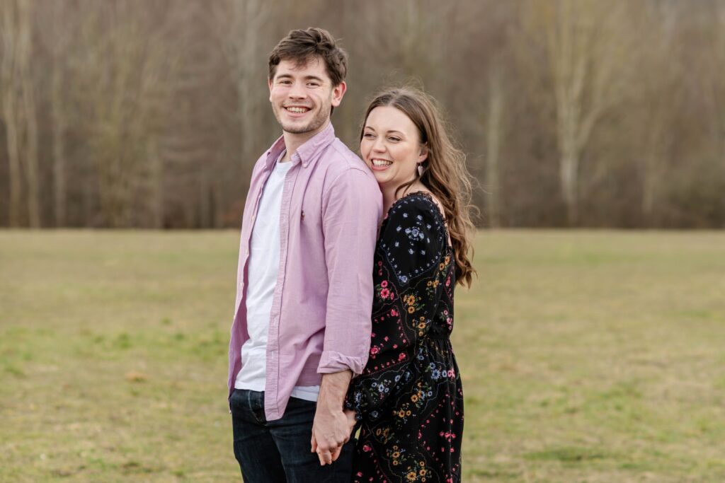 A happy couple holding hands and smiling in a grassy field, with trees in the background.