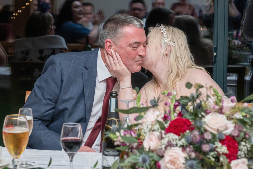 A couple sharing a sweet kiss during a festive gathering, surrounded by flowers and drinks.