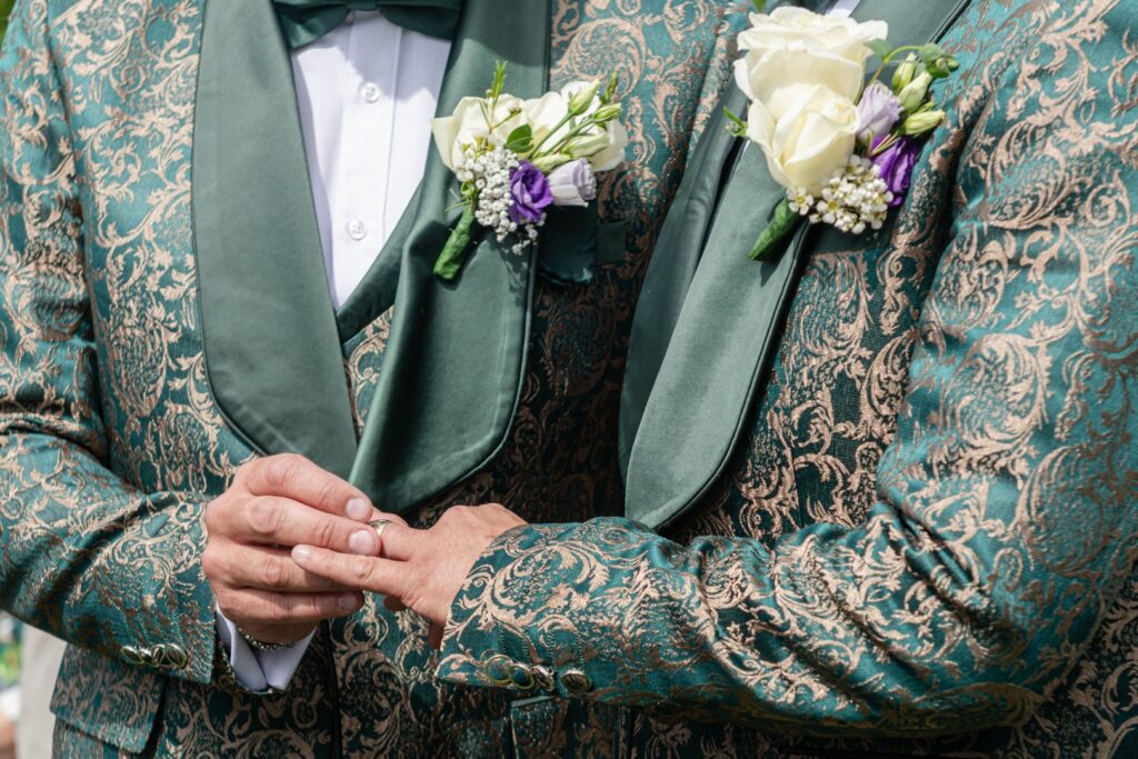Close-up of grooms exchanging rings, showcasing intricate green and gold patterned suits with floral boutonnieres.