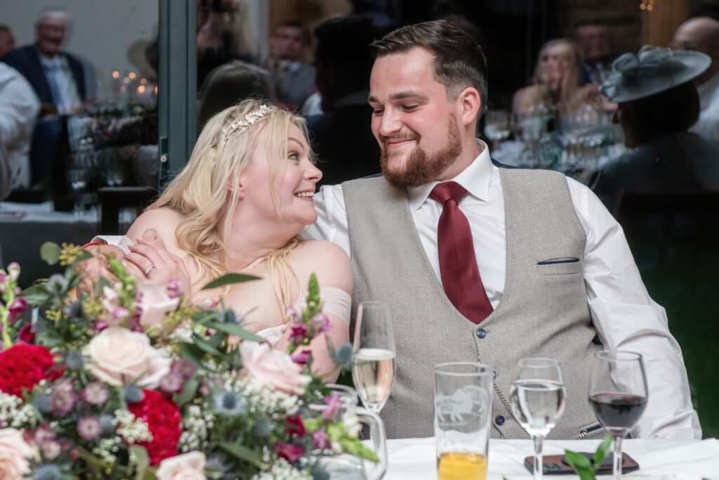 A couple sharing a tender smile during their wedding reception, surrounded by floral arrangements and guests in the background.