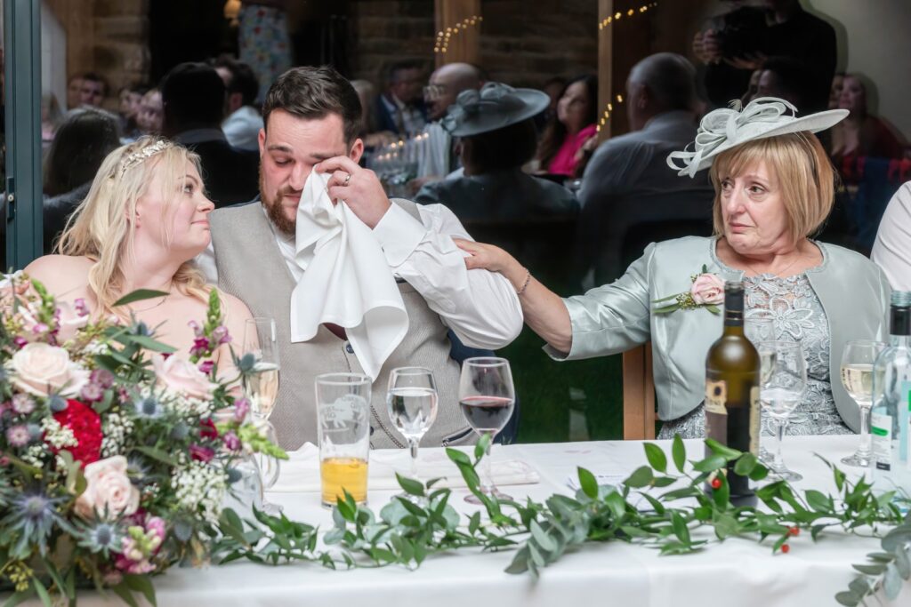 A groom wipes away tears while being comforted by loved ones at a wedding reception table, surrounded by floral decorations and guests in the background.