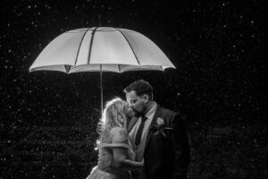 A couple shares a kiss under a white umbrella in the rain, captured in black and white photography, creating a romantic and intimate atmosphere.