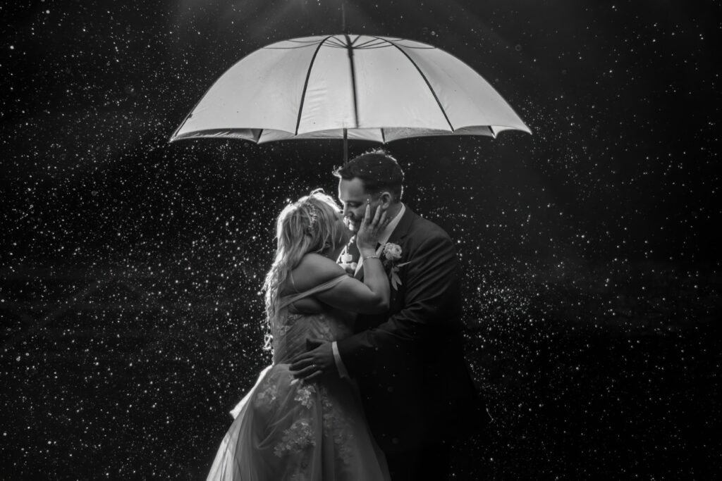 A couple kisses under a white umbrella amid a backdrop of softly falling rain, captured in black and white.