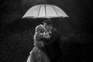 A couple kisses under a white umbrella amid a backdrop of softly falling rain, captured in black and white.