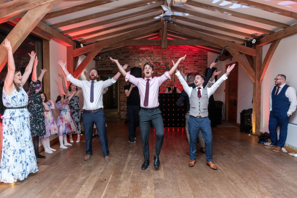 Group of people joyfully dancing with raised arms in a festive indoor setting featuring wooden beams and colorful lighting.