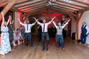 Group of people joyfully dancing with raised arms in a festive indoor setting featuring wooden beams and colorful lighting.