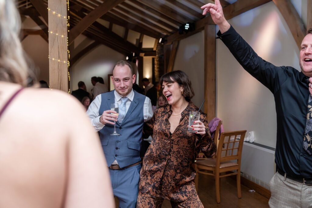 A woman in stylish attire enjoys a drink while dancing with a man in formal wear, surrounded by friends in a festive atmosphere.