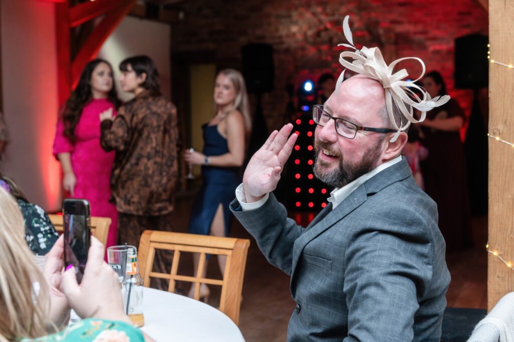 A man in a stylish gray suit and a decorative headpiece smiles and waves while seated at a table, with party guests chatting in the background.