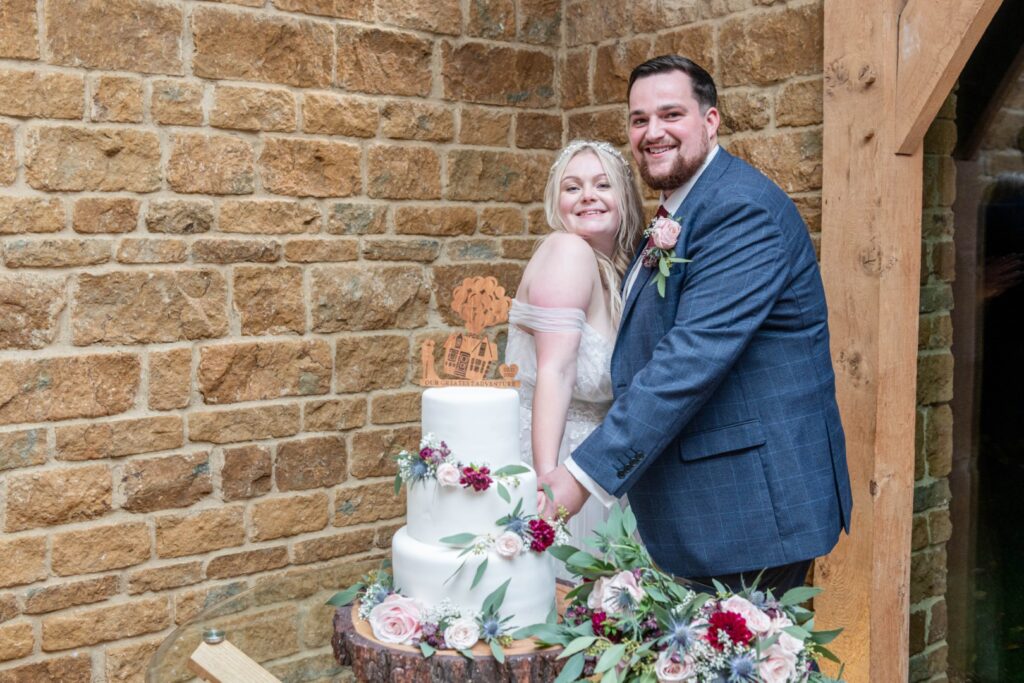 A joyful couple smiles together while cutting their elegantly decorated wedding cake, set against a rustic stone wall.