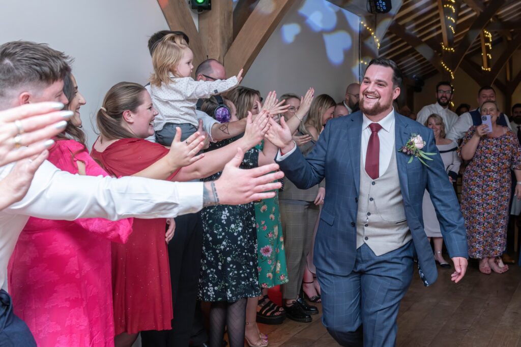 A joyful groom walks through a cheering crowd of guests at a wedding reception, surrounded by smiling faces and raised hands.