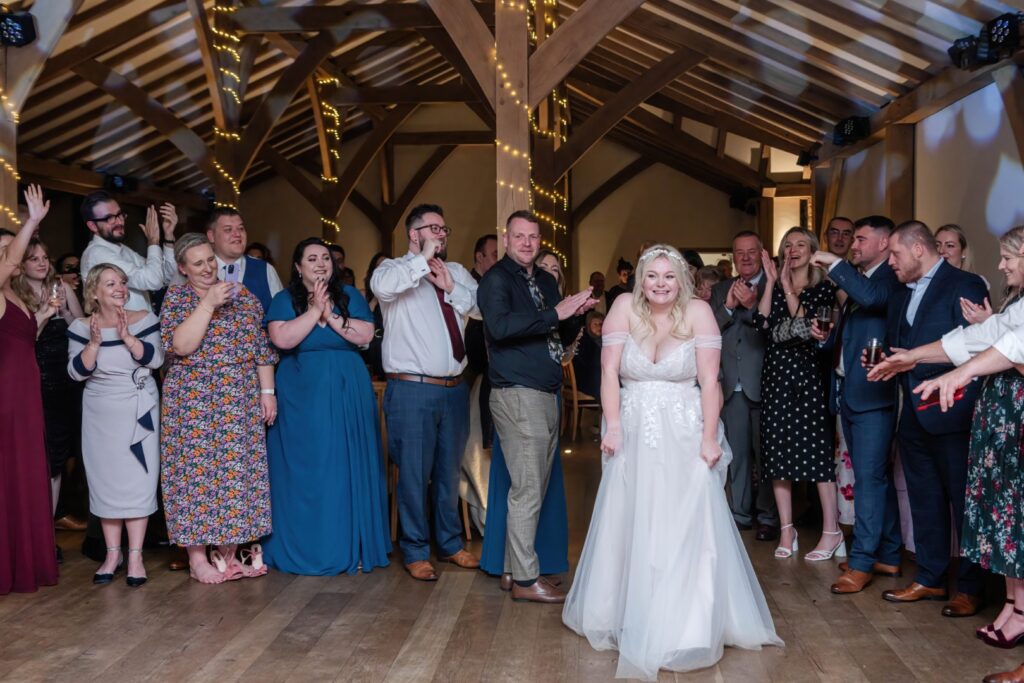 A bride smiles joyfully as she walks towards guests cheering and clapping in a beautifully decorated venue.