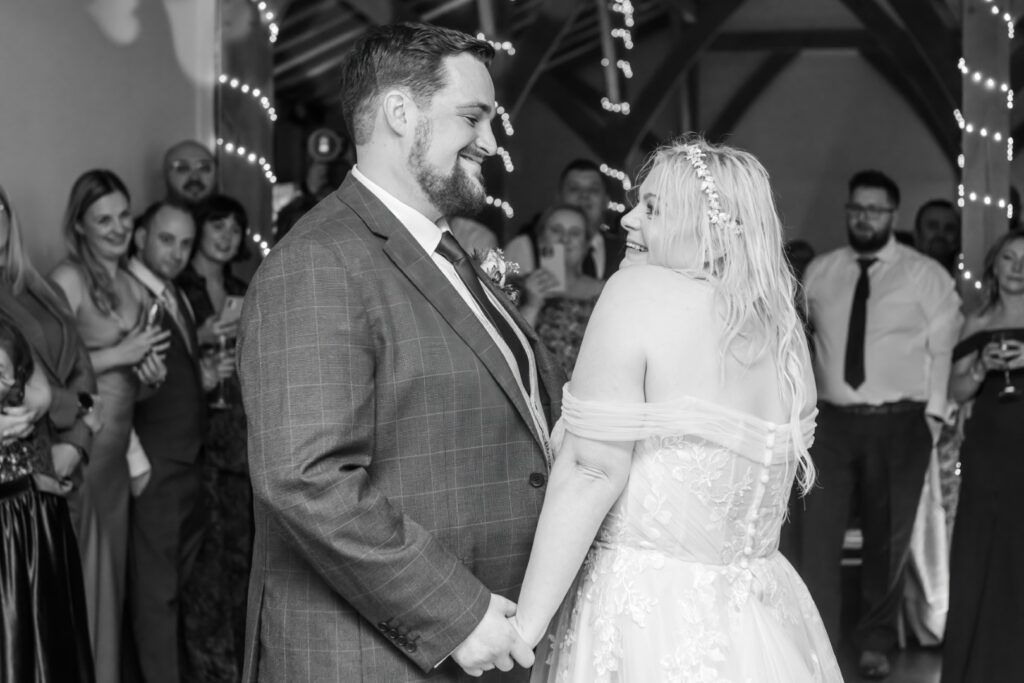 A couple holds hands and smiles at each other during their wedding reception, surrounded by smiling guests.
