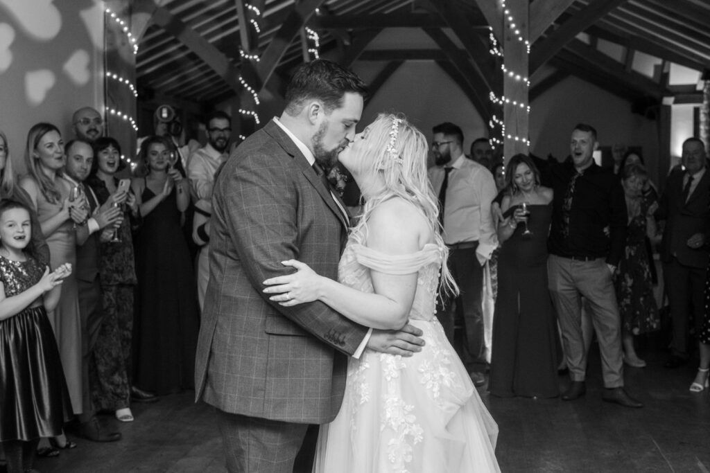 A couple sharing a romantic kiss during their wedding dance, surrounded by an attentive audience in a beautifully decorated venue.