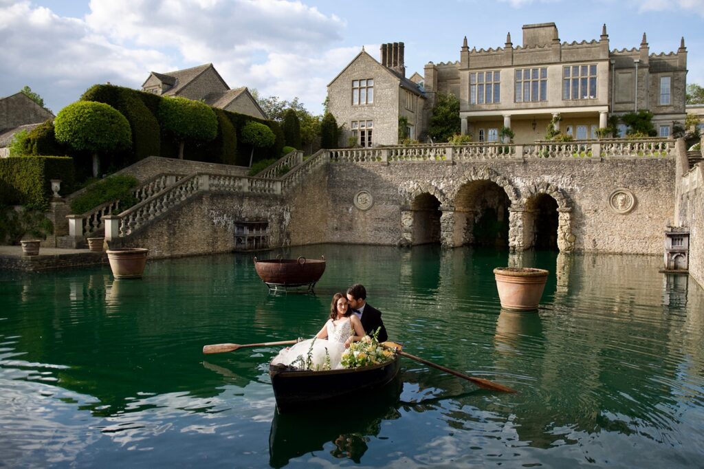 Bride and groom in a rowboat on a turquoise lake in front of Euridge Manor, a fairytale wedding venue in the Cotswolds.