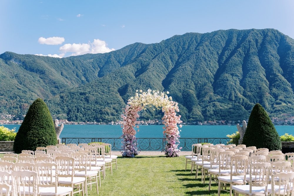 An elegant outdoor wedding setup featuring white chairs, a floral arch, and mountainous landscape by the lake.