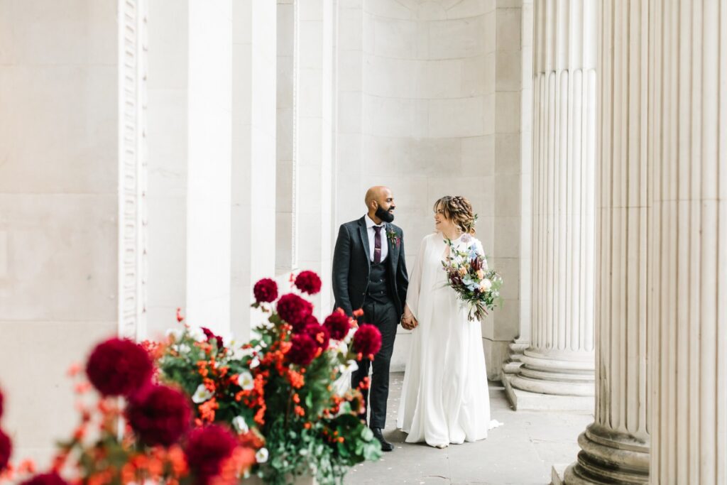 Couple walking hand-in-hand past grand pillars at The Old Marylebone Town Hall, a classic city wedding venue in London.