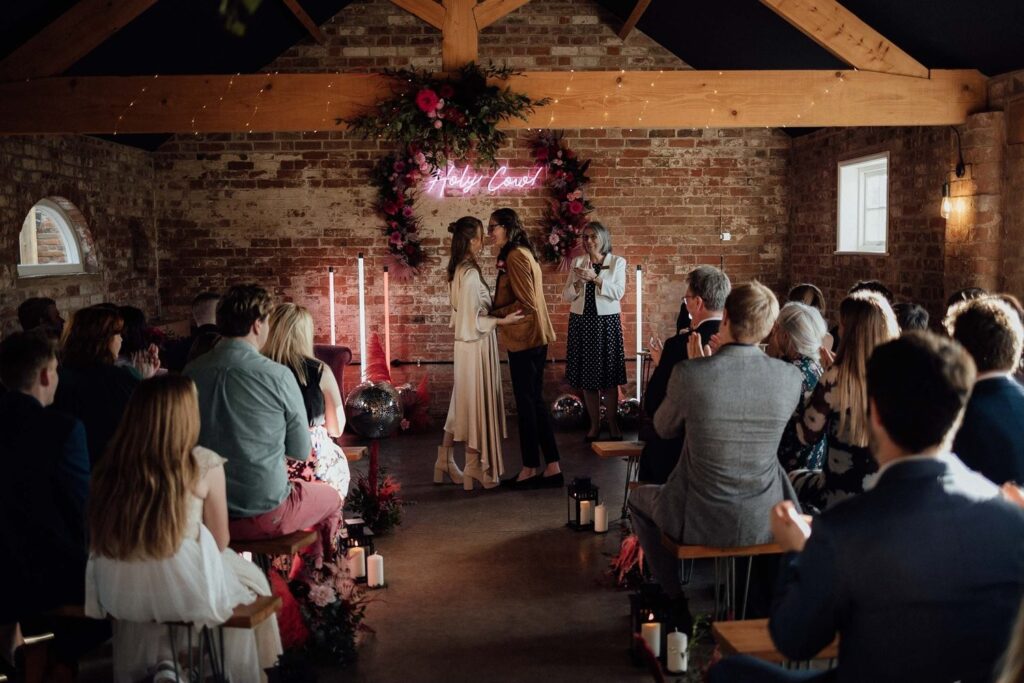 Intimate wedding ceremony kiss under neon sign and floral arch at The Old Cow Shed, a rustic and relaxed barn venue.
