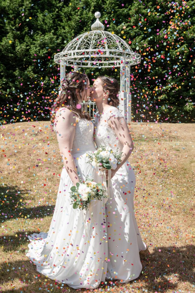 Two brides sharing a kiss amidst a shower of colorful confetti at their outdoor wedding ceremony.