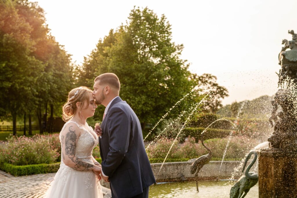 A bride and groom share a tender moment in a garden setting, surrounded by greenery and a fountain in the background.