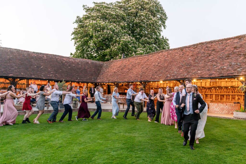 A lively group of people dancing in a line on a grassy area with a rustic barn in the background during a joyful outdoor event.