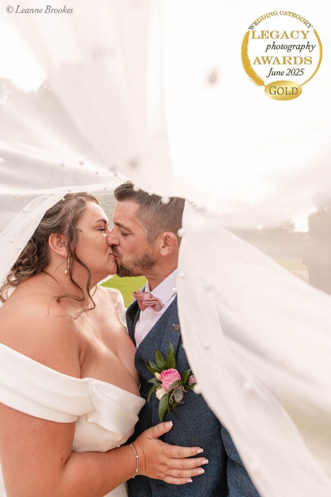 A couple shares a romantic kiss under a delicate wedding veil, surrounded by nature, captured in golden hour light.