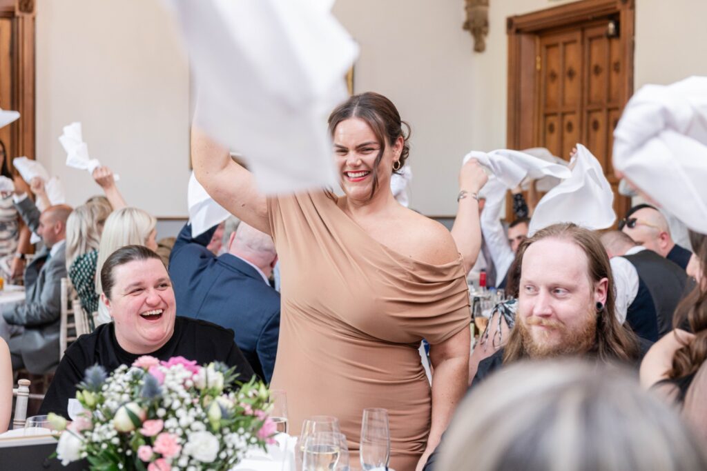 A joyful woman in a brown dress raises a napkin amidst a festive crowd, with attendees smiling and participating in the celebration.