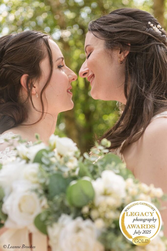 Two brides sharing a tender moment, smiling while holding a bouquet of white flowers in a lush outdoor setting.