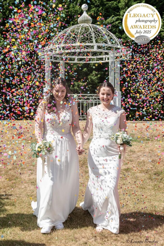 Two brides joyfully walk hand-in-hand through a colorful confetti shower, surrounded by lush greenery and a decorative gazebo in the background.