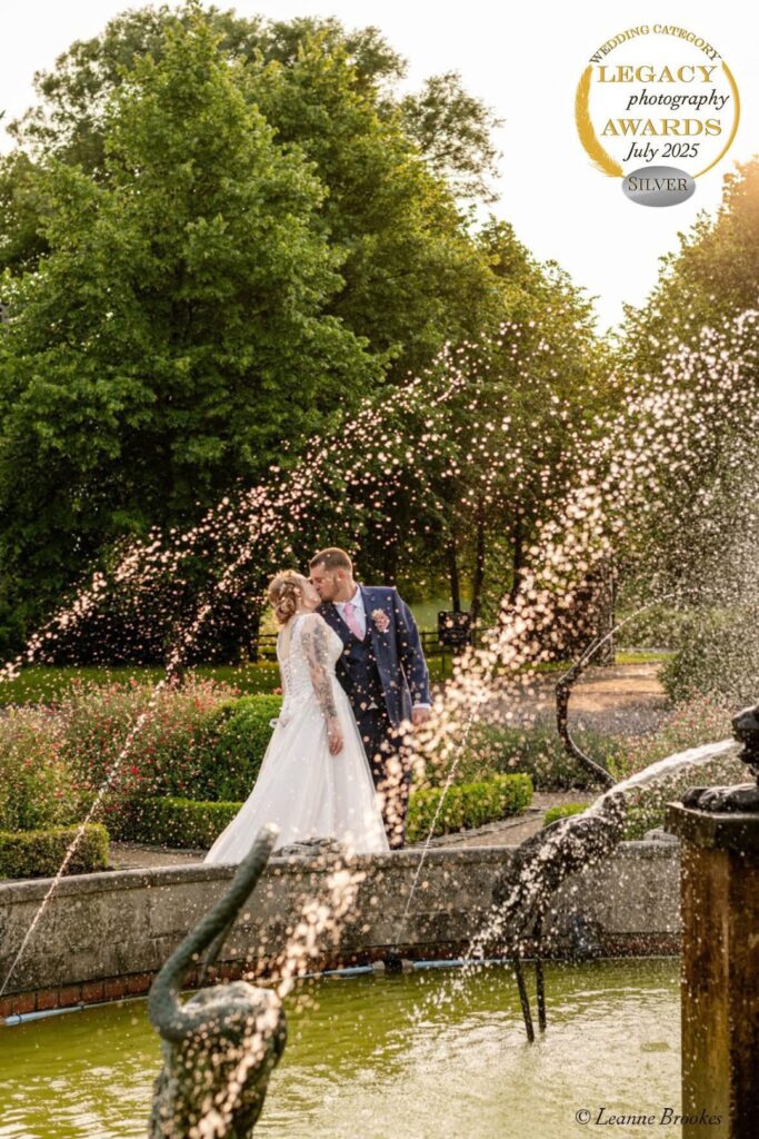 A couple shares a romantic kiss by a fountain amidst beautiful greenery, celebrating their love on a sunny day.