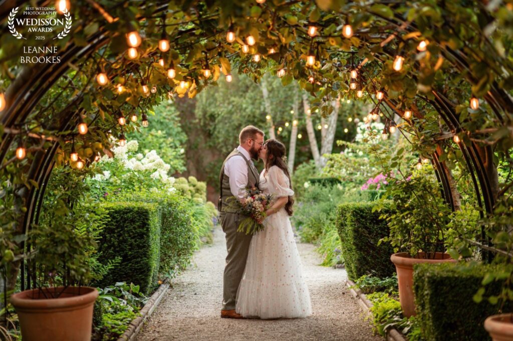 Wedding Photography Portfolio - A couple shares a kiss under a beautifully lit garden arch, surrounded by lush greenery and flowers, capturing a moment of love and intimacy.