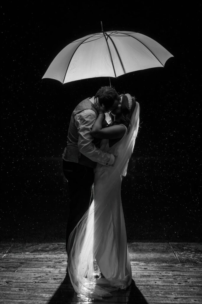 Bride and groom kissing under a white umbrella at night, surrounded by raindrops — a romantic example of rain on your wedding day photos captured by LB Photography.
